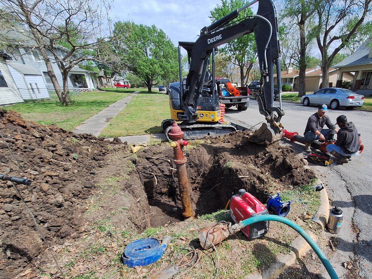 a large hole has been dug with an old hydrant being drained to replace, workers around it