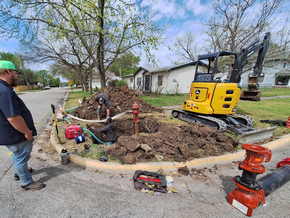 a large hole has been dug with an old hydrant being drained to replace, workers around it