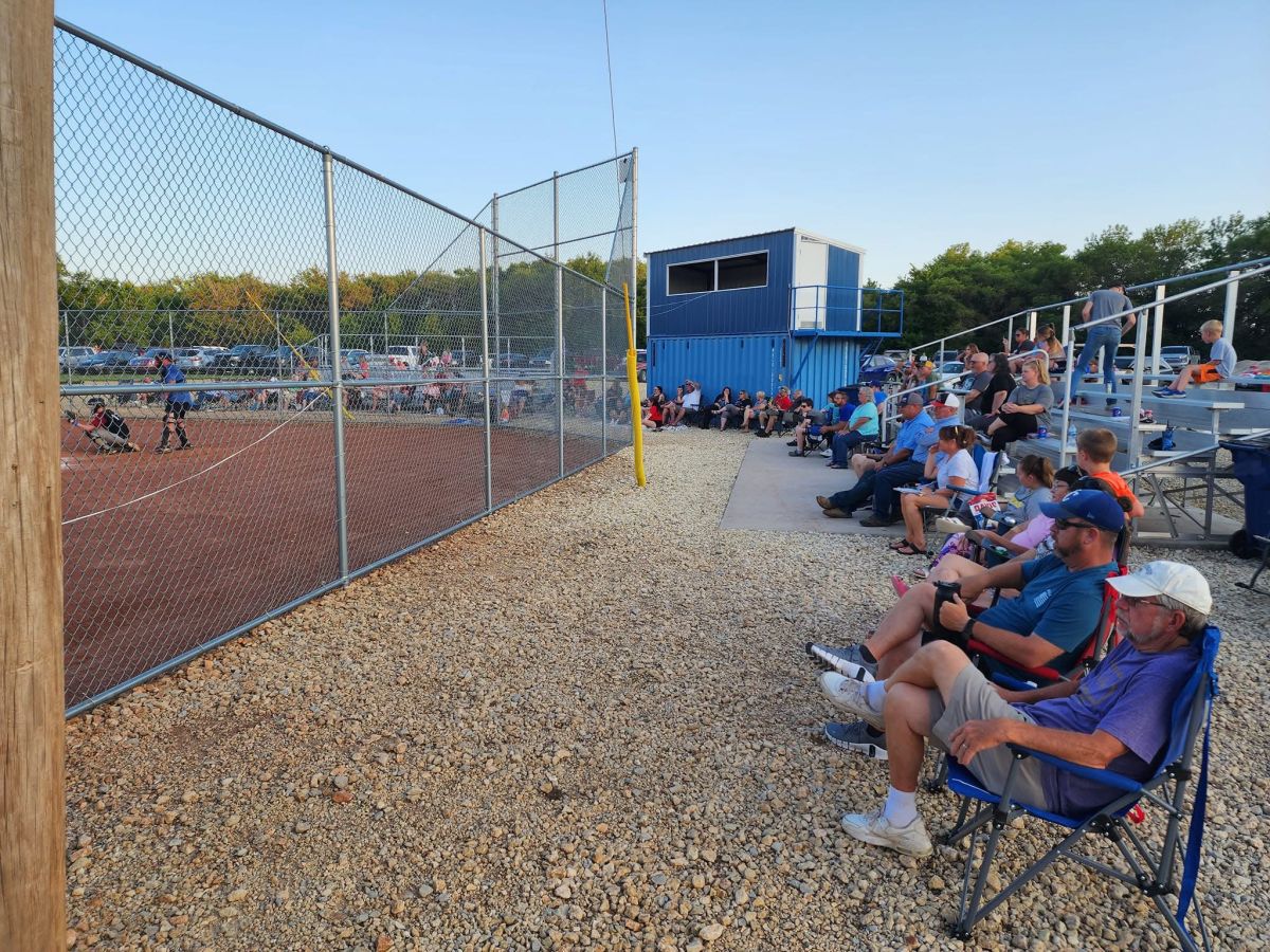people sitting in chairs and bleachers with a press box at a baseball field