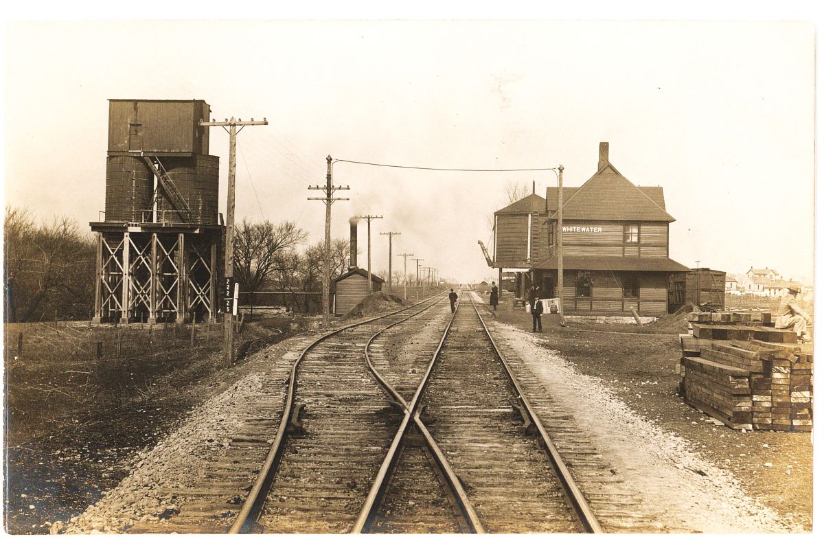 vintage postcard of an old Whitewater Railway station with folks standing on or near the tracks with buildings to the right and water tanks to the left