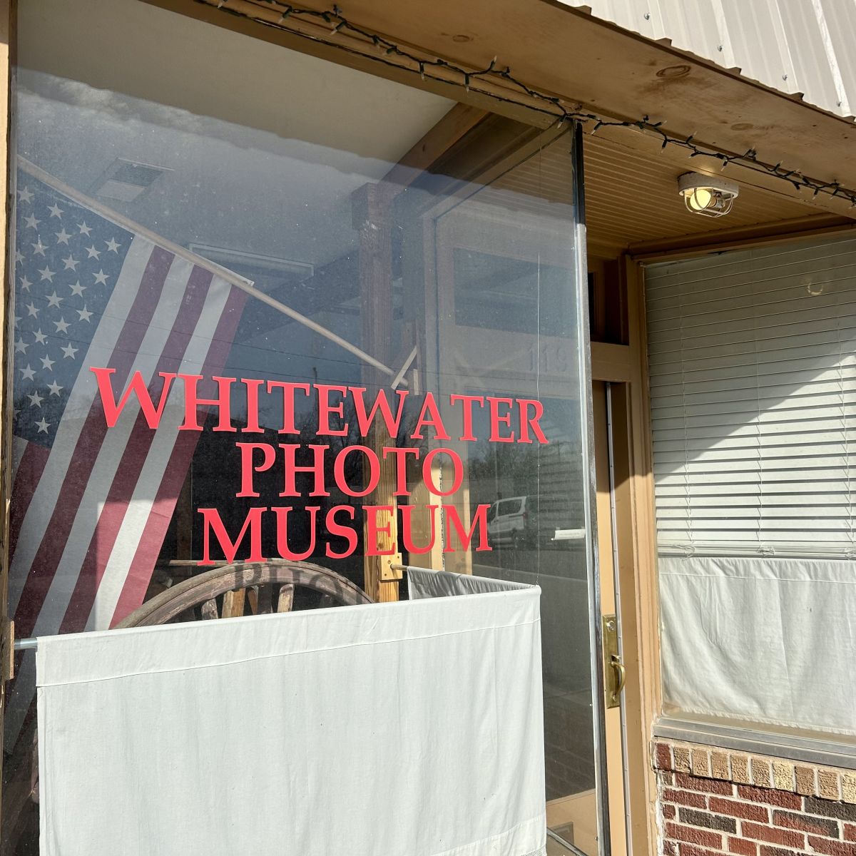 Store front window lettering in red reading Whitewater Photo Museum, inside the window is an American flag and old wagaon wheel